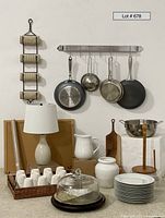 Overall view of the pot rack mounted on a wall holding cookware, mugs, dishes, lamp and other kitchen items visible on floor and wall below