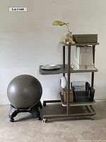 Photo shows the space-saving workstation with shelves laden with various storage boxes, a brass lamp on the top shelf, white folded kitchenware with plates on fold-out side table, and the yoga ball with rolling base beside it.