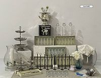 Champagne bucket, flutes, toasting glasses, stoppered bottles, tiered trays, cake server, place card holders, metal table stands, blue vase with flowers, candle holder, salad bowls, tablecloths and runner arranged on white fabric