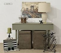 Full lot shot showing small green table on wheels, two storage ottomans, cityscape artwork, lamp, candle and holder, iron jack, reed grass wine carrier with glasses, and metal wine bottle holder with birds.