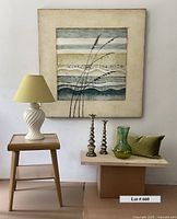View of the mid-century modern blonde wood side table, large canvas artwork in blues and greens with grass, white ceramic lamp with green shade, metal candlesticks, green velvet pillow, and ombré glass vase all arranged together