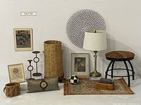 Multiple items displayed on a white wall background including framed artworks, wicker baskets, candle holders, a small rug and stool, antique brass lamp and circular wall decor.