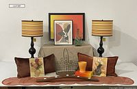 Photo of the full lot with two matching lamps with striped yellow, orange, brown shades, framed artworks, pillows, orange ceramic planter, wire basket and green glass porron decanter on display