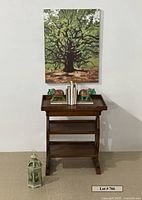 Front view showing wooden side table with three shelves, drawer, elephant bookends holding books, Tree of Life art canvas above, and green metal lantern on floor