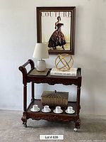Full view of wood carved tea/bar cart with decor items including lamp, gold sphere, book, wooden box, and espresso cups under framed Couture artwork.