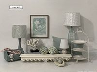 Wide front view of the lot showing lamps, wall shelf with wave motif, coral decor, tiered plate stand with plates, teal wooden trug basket, necklace, and pillows.
