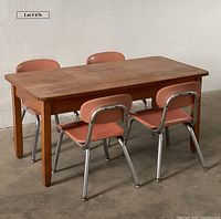 Wide view of four coral Melsur school chairs arranged around a rectangular wooden table on a concrete floor against a white wall.