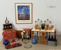 Full display of the lot including framed music festival poster on wall, art glass sculpture, blue tray, metal flower pots, candelabras, pillows, martini glasses, ceramic pitchers, blue bottles in carrier, small table, and other decorative items.