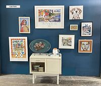 Full view of the white cabinet with drawer and door, surrounded by various sizes of framed art on a blue wall. Painted floral tray displayed on top with mug, candle, and books.