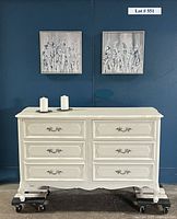 Front view of creamy white, French Provincial style dresser with six drawers, ornate handles, and scalloped base, two canvas floral prints on blue wall behind, two white candles on top of dresser.