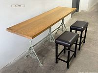 View of the homemade work table showing thick wooden top with scratches and three cast iron legs with scrollwork. Two black vinyl padded stools placed beside it.
