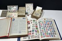 Two hardcover albums open displaying mounted and loose stamps, boxes and envelopes of loose stamps scattered on table