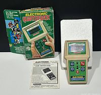 Photo showing the game, original box, and instruction manual on a black background. The box is green with football player graphics and game features listed. The handheld game has a green control panel with buttons and a display screen.
