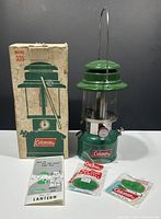 Photo of vintage Coleman 335 Sport-Lite lantern displayed with original box, manual, and unopened mantles on white surface against dark background.