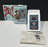 Photo showing the handheld game in original foam packaging, cardboard box with hockey players and branding, and instruction manual laid out on a white surface.