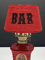 Front view of the vintage kerosene lamp showing the red shade with 'BAR' text and lenticular nude image on the porcelain base.