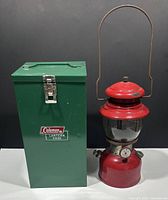 Photo showing the Coleman red lantern beside the green metal carrying box, showcasing the full set.