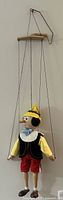 Full view of Pinocchio marionette hanging from wooden control bar with strings attached to limbs and head.