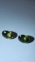 Pair of oval cut tricolored sapphires showing yellow, green, and blue undertones lying side by side on a flat surface.