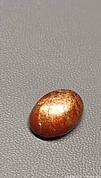 Oval natural sunstone cabochon shown on a textured dark surface highlighting its coppery-orange metallic flash.
