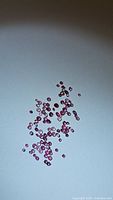 Top view of multiple small round diamond cut pink sapphires scattered on a white background with varying shades of pink and red, size ranges about 1-1.5mm.