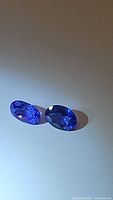 Two faceted oval tanzanite gemstones placed side by side on a light surface, showing deep purple blue color and facets.