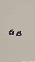 Pair of natural purple amethyst gemstones in trillion cut shape, positioned on neutral background, showing front and side views to display cut and clarity.
