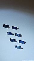 Seven natural blue sapphires, baguette cut, arranged on a white background showing various angles and lighting revealing deep blue color and facets.