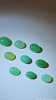 Photo showing nine oval cabochon green chrysoprase gemstones arranged on white background with natural and translucent variations in green shades.