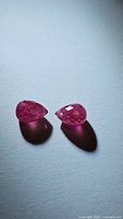Two pink-red faceted briolette cut rubies showing teardrop shape and drilled holes for jewelry attachment, on white surface under soft directional lighting.