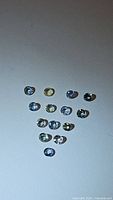 Top down view of fifteen small round faceted pastel sapphires arranged on a white surface showcasing subtle variation in colors including blue, yellow, green, and violet.
