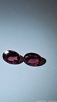 Two oval-shaped pink-purple rhodolite garnet gemstones side by side on a white surface, showing their color and clarity.