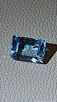 Close-up image of natural Swiss blue topaz gemstone, baguette cut, showing clarity and blue lagoon color on textured background.