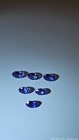 Six natural oval cut tanzanite gemstones, blue-purple in color, laid flat on a white surface under light, showing clarity and color consistency.