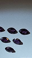 Six natural purple marquis cut sapphire gemstones arranged on a flat surface with a soft shadow visible, showing their faceted cuts and rich purple color.