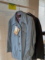 Two men's leather jackets hanging on a rack by a window with blinds. Light blue Sears The Boulevard Club jacket in front, darker jacket behind.
