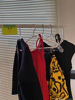 Five vintage dresses hanging on plastic hangers on a curtain rod near window blinds, including a red dress, yellow patterned dress, and dark dresses