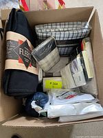 Box containing black fleece throw blanket and multiple bed linens including pillow protectors and sheets, all varied sizes, mostly in packaging.