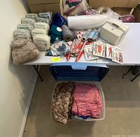 Wide view of table and box containing crafting supplies: wool packs, fabric, patterns, knitting needles, and sewing accessories.