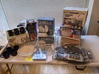Photo of kitchenware items on table including boxed Corelle dishes, boxed CorningWare Rangetop cookware set, slow cooker, black and clear mugs, cooking utensils and glassware.