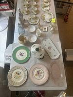 Wide view of assorted fine china items including teacups, saucers, plates, glass dish, and figurines arranged on a table.