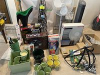 Wide view of the garden and household lot including various green and yellow plant pots, boxed outdoor security light, black metal cage holder, solar garden light, insect zapper light, and part of oscillating fan box.