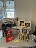 Full view of boxed kitchen items on table including covered paella pan, boxed Pomona glasses, T-fal pressure cooker, ceramic mugs, onion baker, and kitchen towel set.