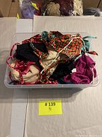 Clear bin container holding multiple assorted vintage women’s swimsuit pieces in various colors and patterns, mostly tops visible