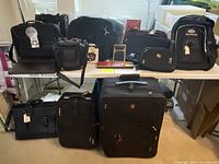 Photo showing entire lot of luggage and bags arranged on and under two tables, all mostly black in color.