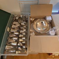 Photo showing silver plated stemware pieces laid inside a plastic container and a punch bowl set in a cardboard box.