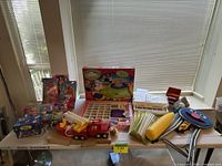 Wide view of assorted toys, craft kits, and boxed Barbies on table with blinds background.