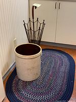 Photo showing the crock, braided rug and metal umbrella stand placed on a tiled floor near a white cabinet.