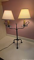 Black wrought iron 2-bulb lamp illuminated, showing beige fabric shades, and tripod base.