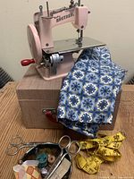 Pink Baby Brother battery-operated sewing machine on top of brown hardcase suitcase with sewing tools and blue patterned fabric